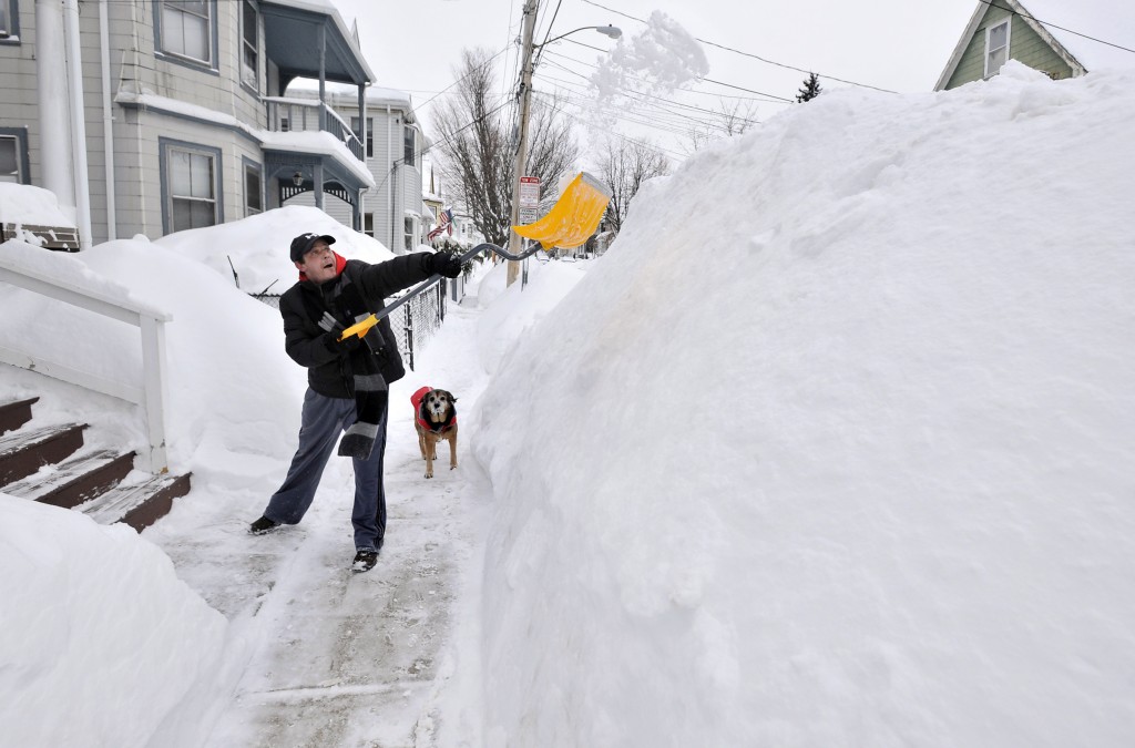 Lee Anderson adds to the pile of snow beside the sidewalk in front of his house in Somerville, Mass., Tuesday, Feb. 10, 2015, as his dog Ace looks on. The latest snowstorm left the Boston area with another two feet of snow and forced the MBTA to suspend all rail service for the day. (AP Photo/Josh Reynolds)