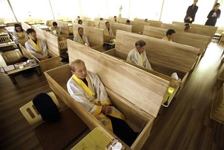 Wang Yong-yo, left bottom, sits inside a wooden coffin during the “death experience” program at Hyowon Healing Center in Seoul, South Korea, Tuesday, Dec. 22, 2015. In a dark, dimly-lit room, people dressed in white burial shrouds sit down next to dozens of coffins. They write their wills, climb into the caskets, lie down and a symbolic “angel of death” _ a man wearing a traditional Korean hat and black robes -- shuts the lid of each casket.(AP Photo/Ahn Young-joon)