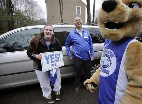 In this Tuesday, March 15, 2016 photo, Maya Wolf, 17, left, holds a sign to her mother, not shown, from the driveway of their home in Franklin, Mass., as she is visited by Wheaton College President Dennis Hanno, center, and the school's mascot. Wolf was presented with an acceptance letter from Wheaton College officials during the visit. More colleges have started to hand-deliver portions of their acceptance letters to students. (AP Photo/Steven Senne)